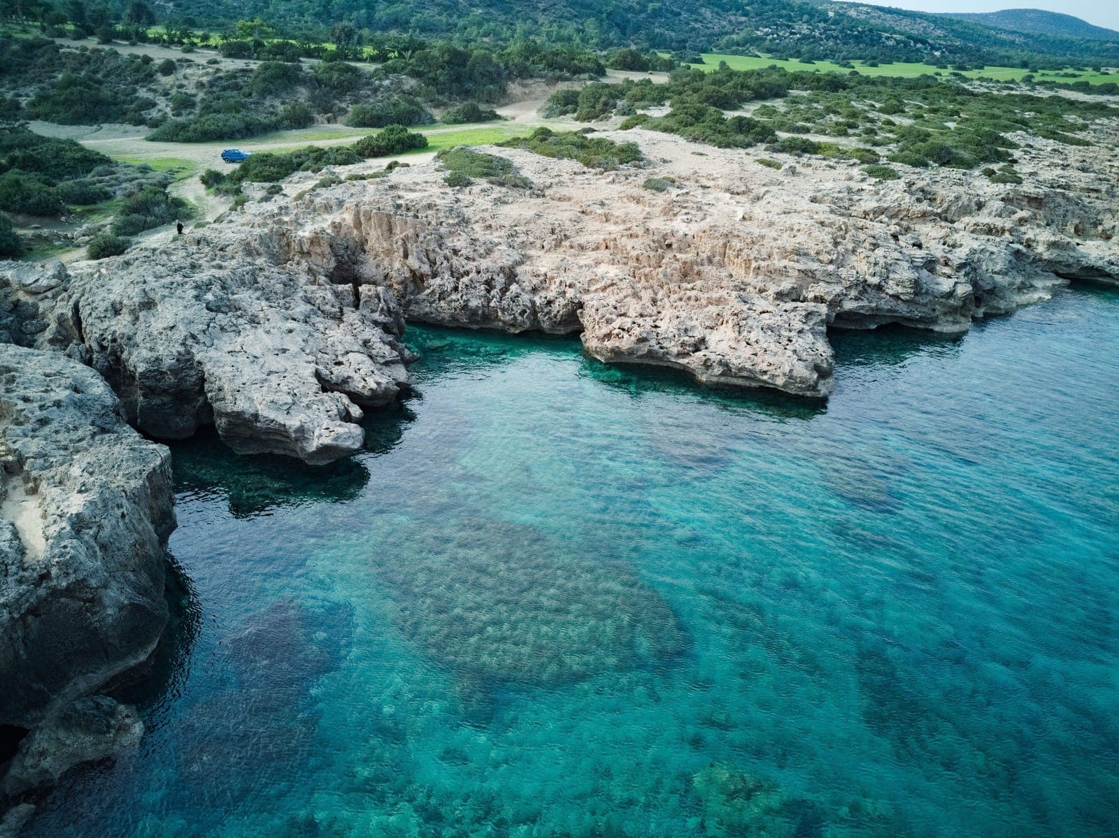 Aerial view of the undeveloped Akamas coastline: jagged limestone rocks over clear turquoise water, with scrubland and a dirt track behind
