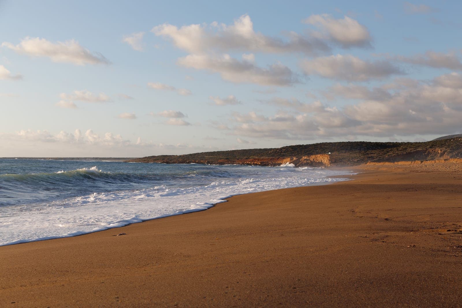 The empty sandy shore of Lara Bay on the Akamas coast, with waves breaking and low scrub-covered headlands behind
