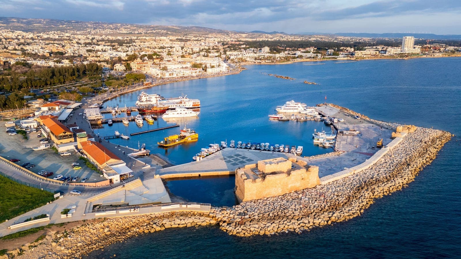 Aerial view of Paphos harbour at golden hour: Paphos Castle on the breakwater, day-cruise boats and fishing boats moored, and the Kato Paphos seafront curving behind