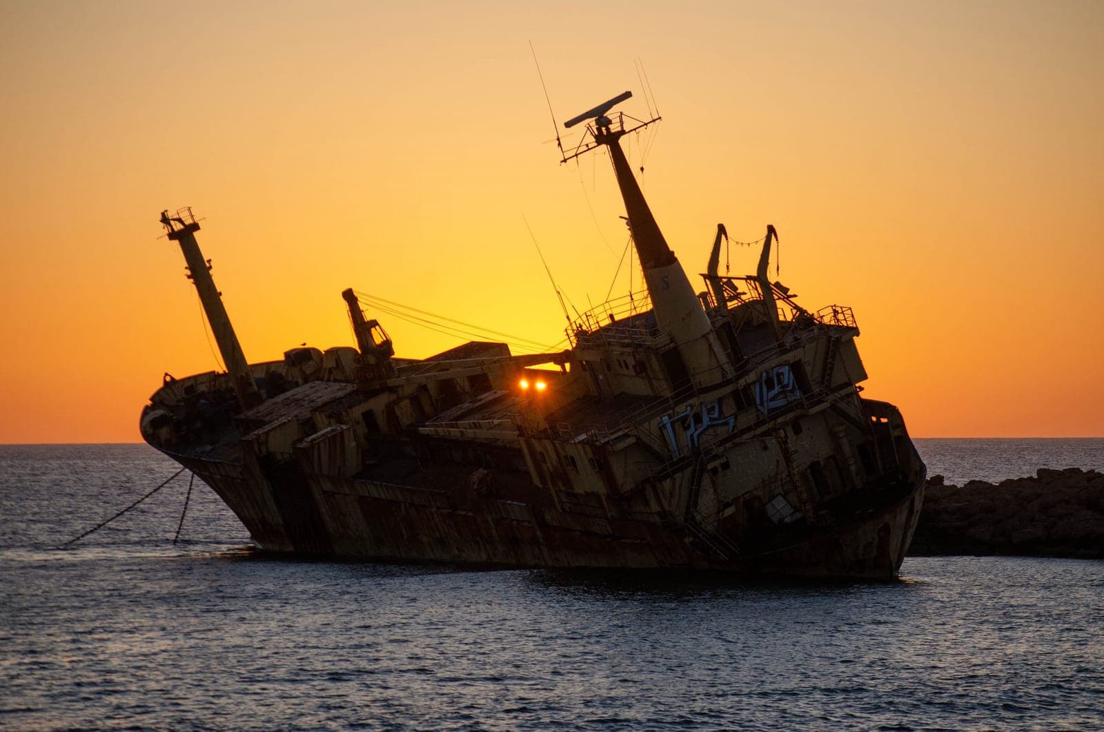 The stranded Edro III cargo ship listing on the rocks off Peyia, silhouetted against an orange sunset sky