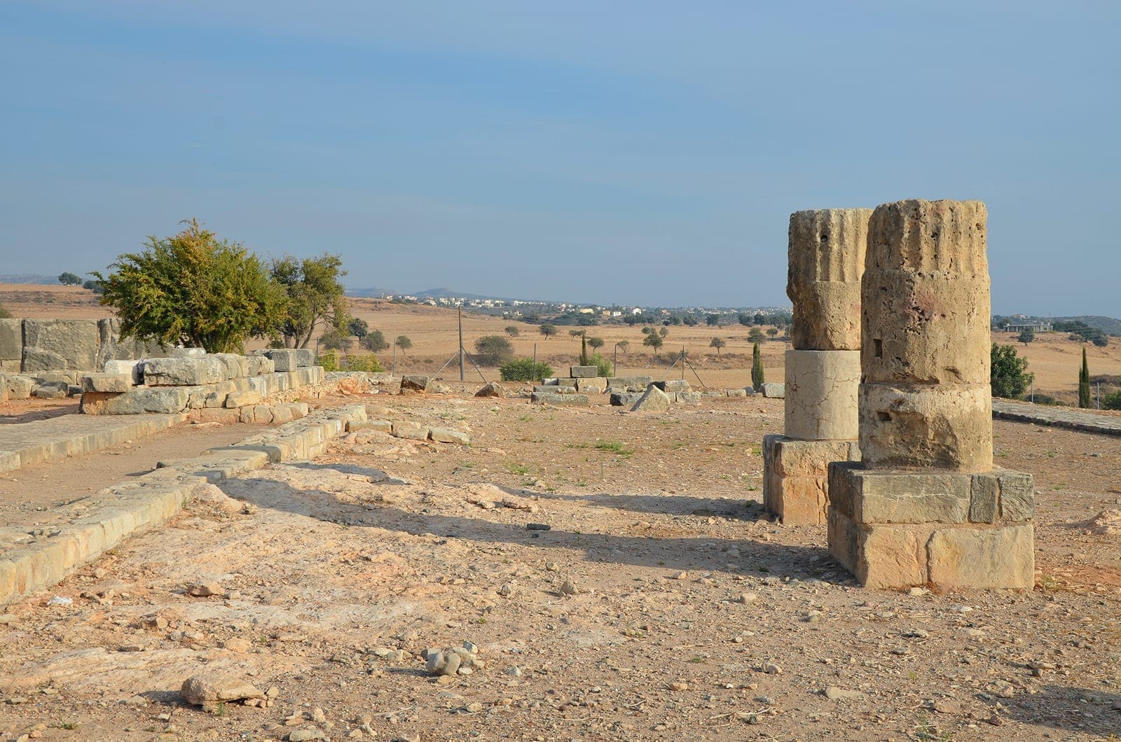 Low stone foundations and two column drums at the Sanctuary of Aphrodite in Kouklia, with dry fields and a village in the distance